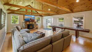 A spacious living room with a wooden ceiling, a fireplace, a pool table, and a large window offering a view of the outdoors.