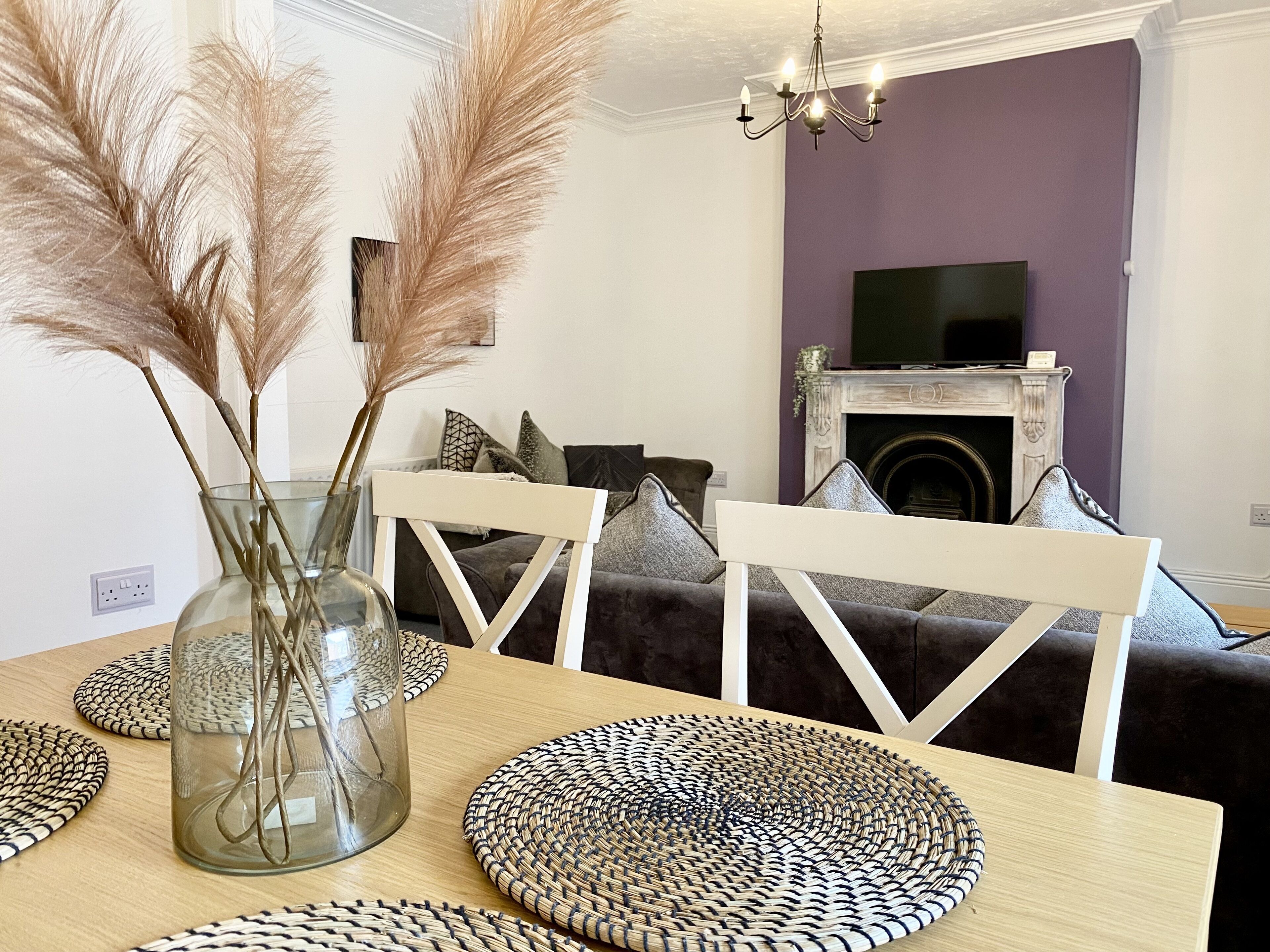 A dining area with a table set for two, featuring woven placemats and a glass vase with pampas grass.