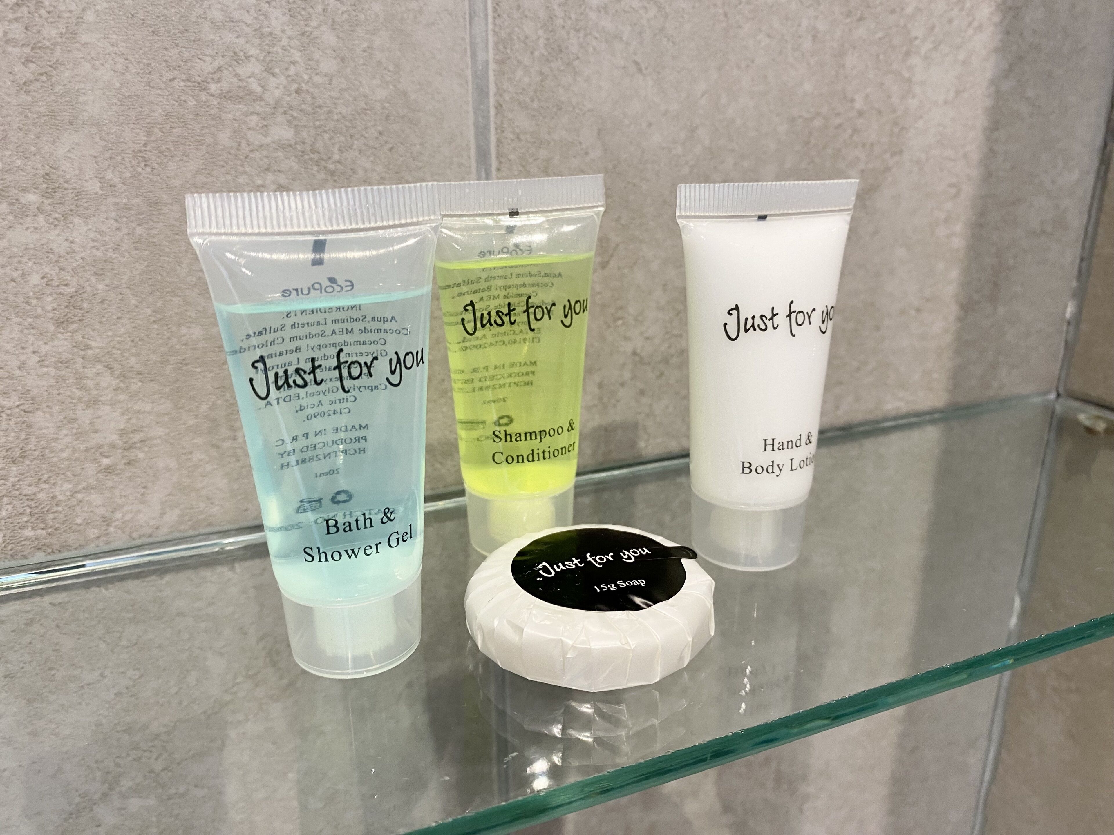 Kylpyhuoneen hyllyllä on kolme hygieniatuotetta: sininen pullo, jossa lukee «Suihkusaippua», keltainen pullo, jossa lukee «Shampoo ja hoitoaine», ja valkoinen pullo, jossa lukee «Käsi- ja vartalovoide».