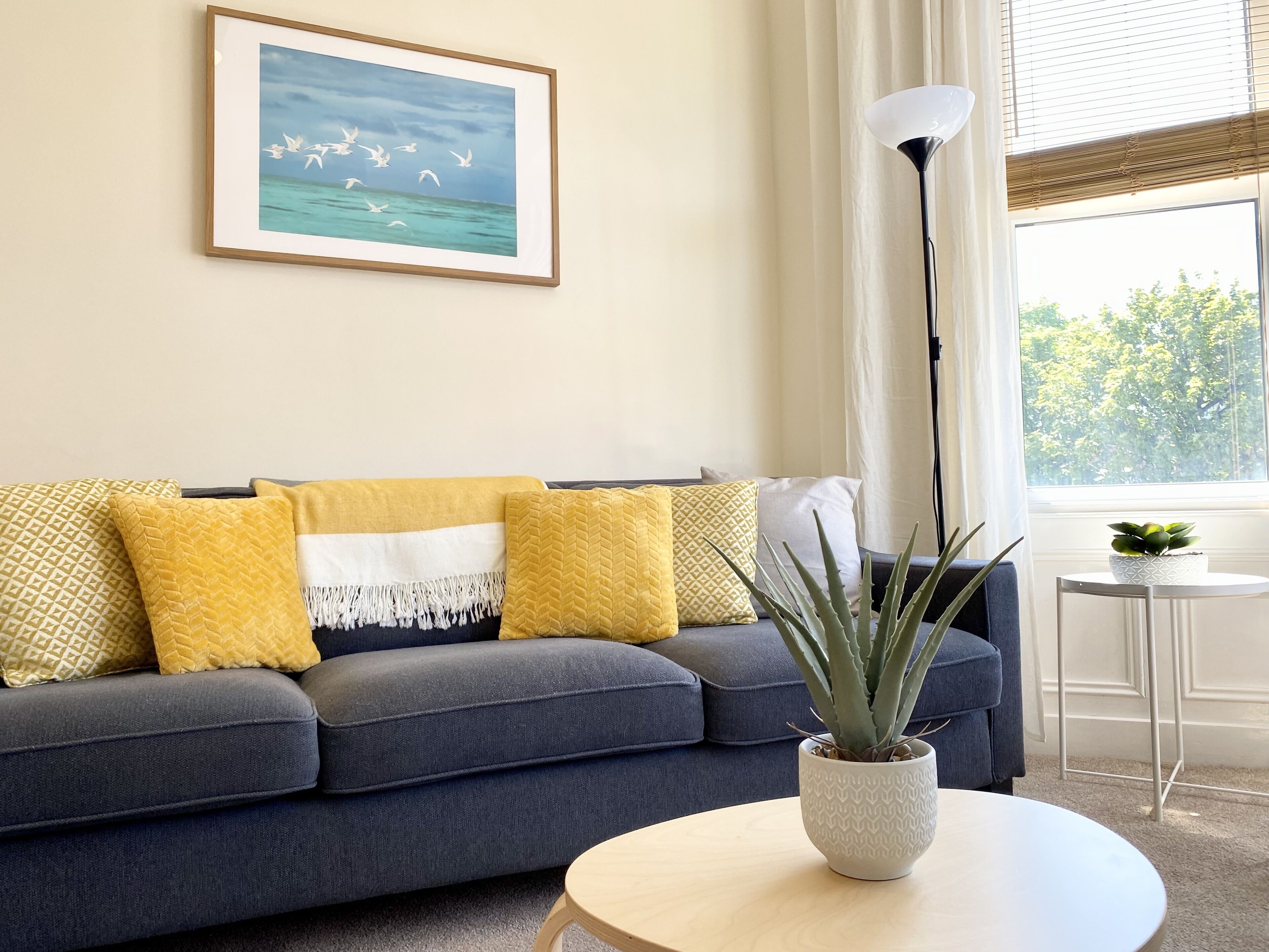 Een woonkamer met een grijze bank, geel-witte kussens, een lijst met een afbeelding van vogels boven water, een hoge vloerlamp en een potplant op een ronde tafel.