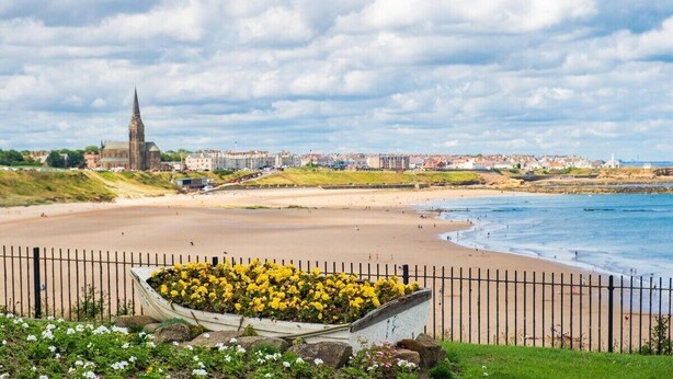 Un pueblo costero con una imponente aguja de iglesia, una playa arenosa y un parterre floral en primer plano.