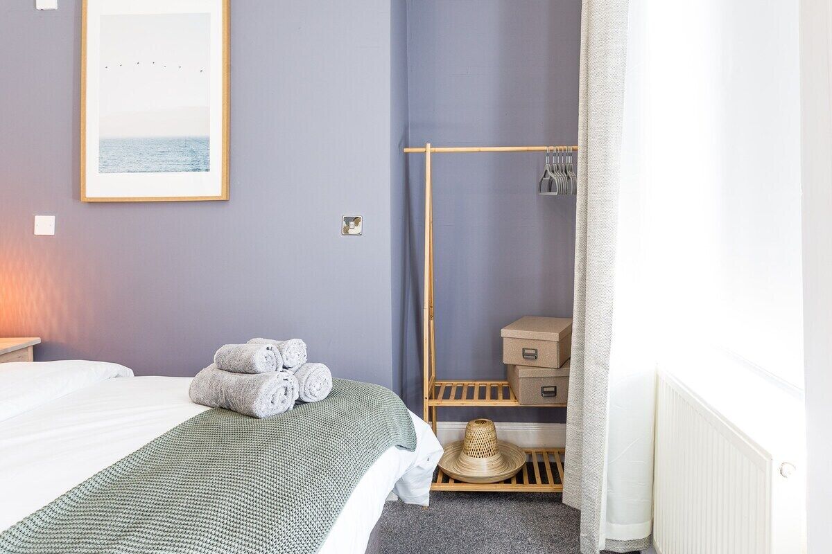 Une chambre d’hôtel bien rangée, avec un lit, une étagère en bois où sont posées des serviettes, un tableau encadré au mur et un radiateur.