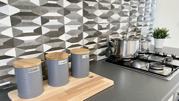 Een keuken met een moderne backsplash met geometrische patronen, een gasfornuis en drie cilindervormige bakjes met de aanduidingen 'koffie', 'suiker' en 'thee' op een houten snijplank.