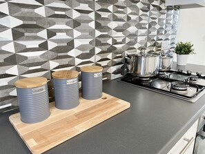Eine Küche mit einer modernen, geometrischen Spritzschutzwand, einem Gasherd und drei zylindrischen Behältern, auf denen 'Kaffee', 'Zucker' und 'Tee' stehen, auf einer Holzschneidebrett.