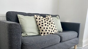 Een grijze bank met twee kussens, waarvan één effen en één met stippen, in een kamer met lichtgrijze vloer.