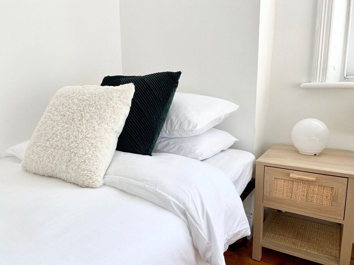Un lit bien fait, avec une literie blanche et deux oreillers, un blanc et un foncé. Une table de chevet en bois, dotée d’un tiroir en osier, se trouve à côté du lit.