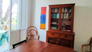 A dining area with a wooden table and chairs, a bookshelf, and a door leading to an outdoor area with a view of trees. - The White House on the sea (Rosignano Marittimo)