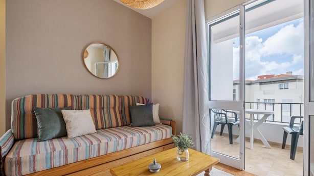 A living room with a striped sofa, a round mirror, a wooden coffee table, and a view of a balcony with outdoor furniture. - Escapadinha do Sol by Homie (Porto Santo)