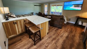 Un salon chaleureux avec un plancher en bois, un canapé en cuir marron, un îlot en marbre blanc avec des tabourets, et un téléviseur à écran plat fixé au mur.