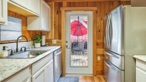 Une cuisine compacte avec des meubles blancs, un réfrigérateur en acier inoxydable, un évier et une porte donnant sur une terrasse extérieure agrémentée d’un parasol rouge.