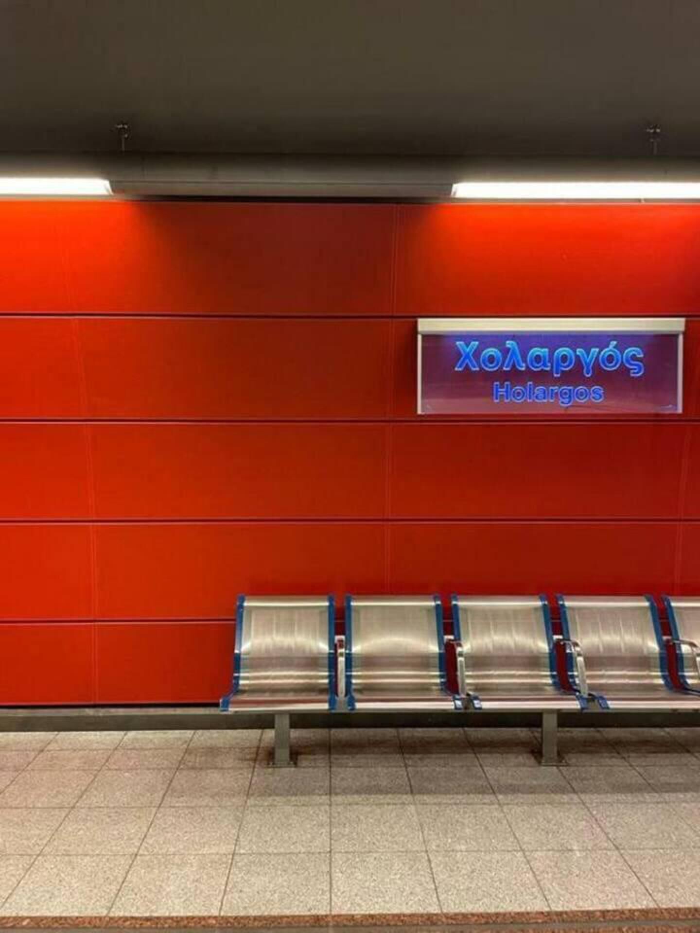Uma área de espera com parede vermelha e um letreiro com a inscrição "Холарюс Холаргос". Há bancos de metal alinhados contra a parede.