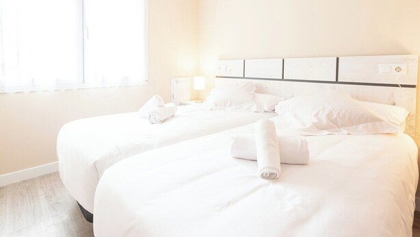 A hotel room with two beds, white bedding, a headboard with a light switch, and a window with sheer curtains.