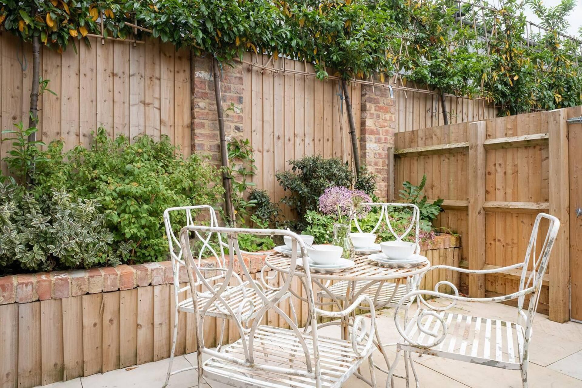 A patio with a metal table and chairs, surrounded by a wooden fence and greenery.