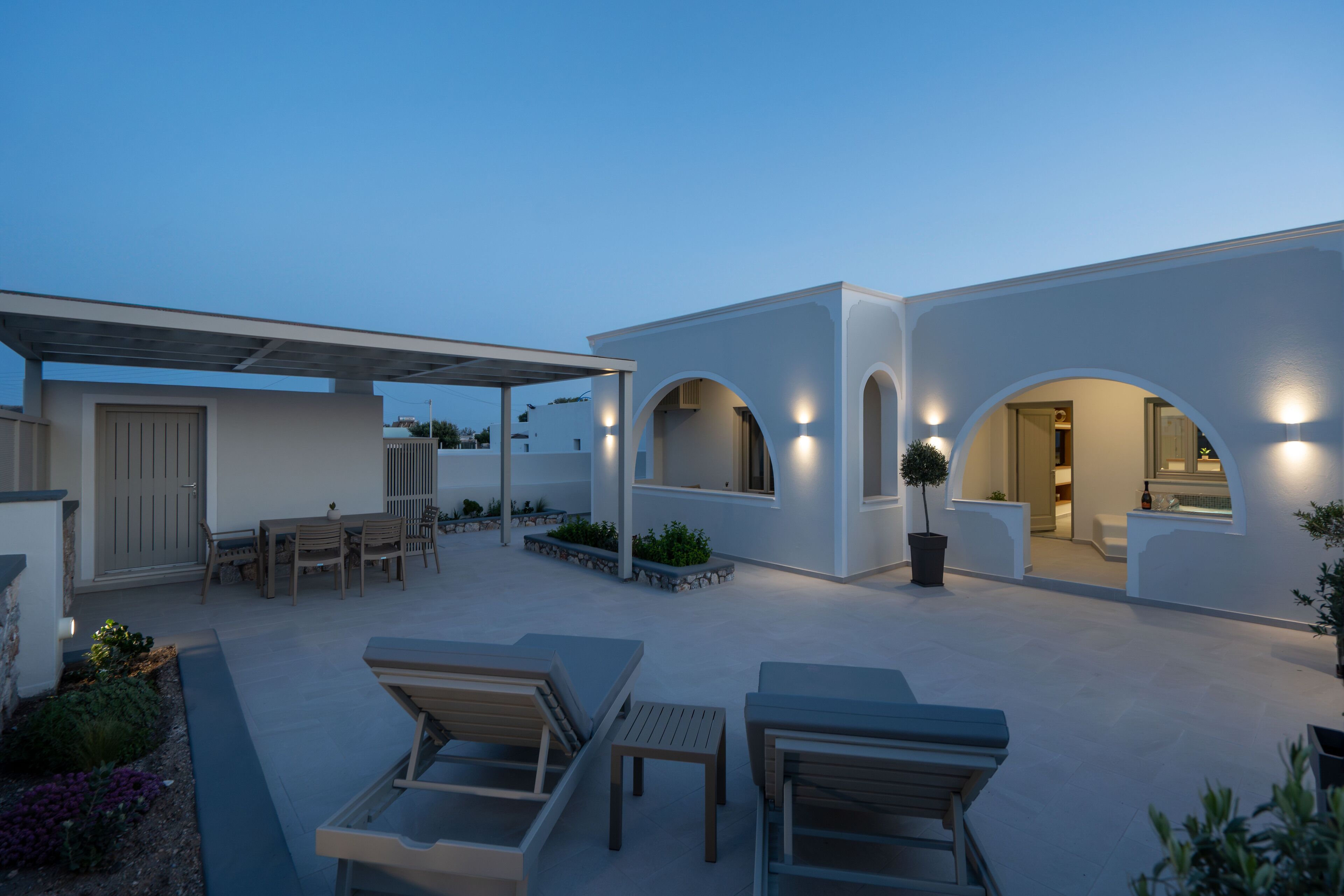 A modern outdoor patio with seating and a covered area, featuring a minimalist architectural design with arched openings and ambient lighting.