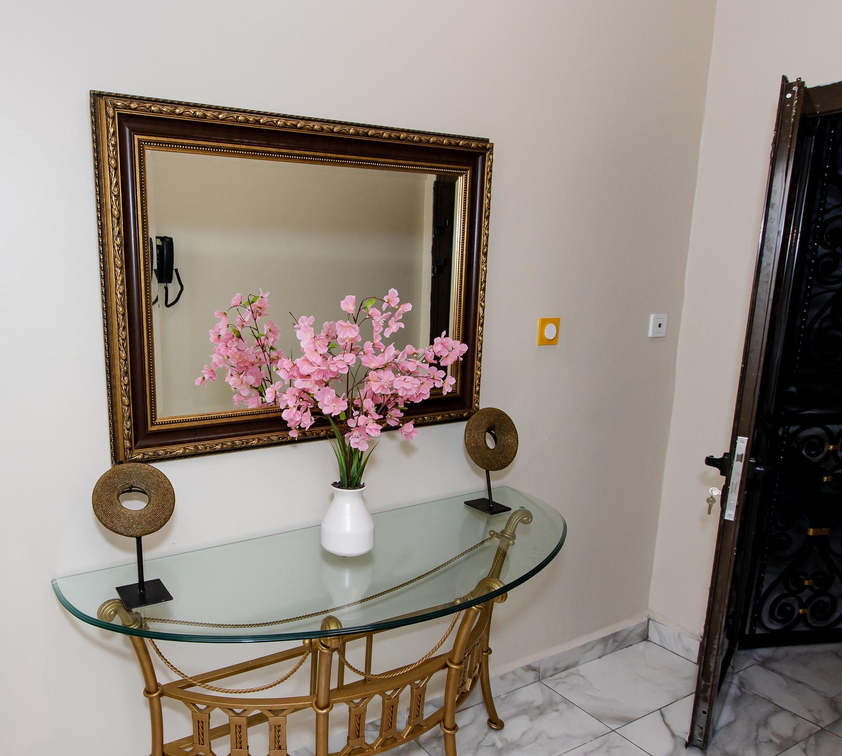 A glass-top desk with a white vase of pink flowers, a gold-framed mirror, and two decorative gold rings on stands.