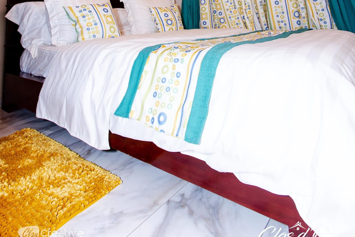 Un dormitorio con techo de madera, cama con ropa blanca y almohadas estampadas, alfombra amarilla y ventana con cortinas azul verdoso.