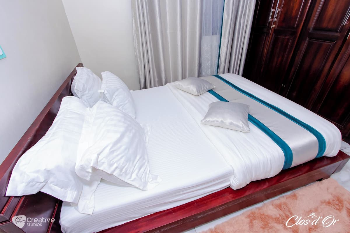 Una cama bien tendida con sábanas blancas y un detalle en color verde azulado, un cabecero de madera y una alfombra rosa y mullida.
