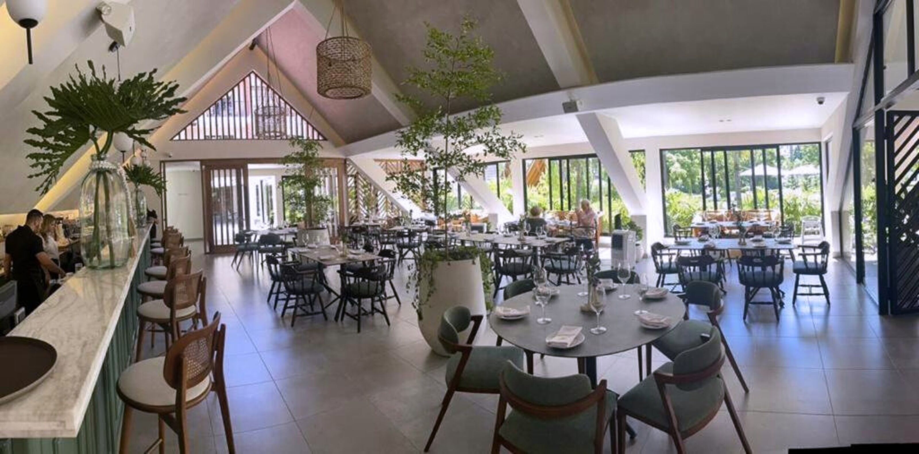 Un restaurante espacioso con techos altos, amplias ventanas y una planta en maceta central. Las mesas están dispuestas con copas de vino y platos.
