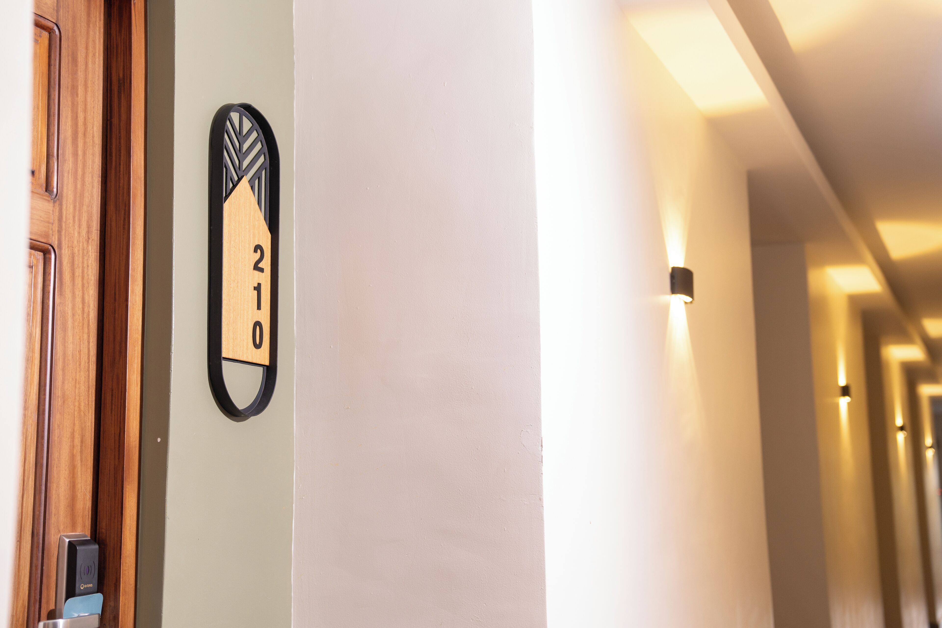 Un couloir d’hôtel avec une porte en bois et une plaque fixée au mur indiquant le numéro de chambre : 210.