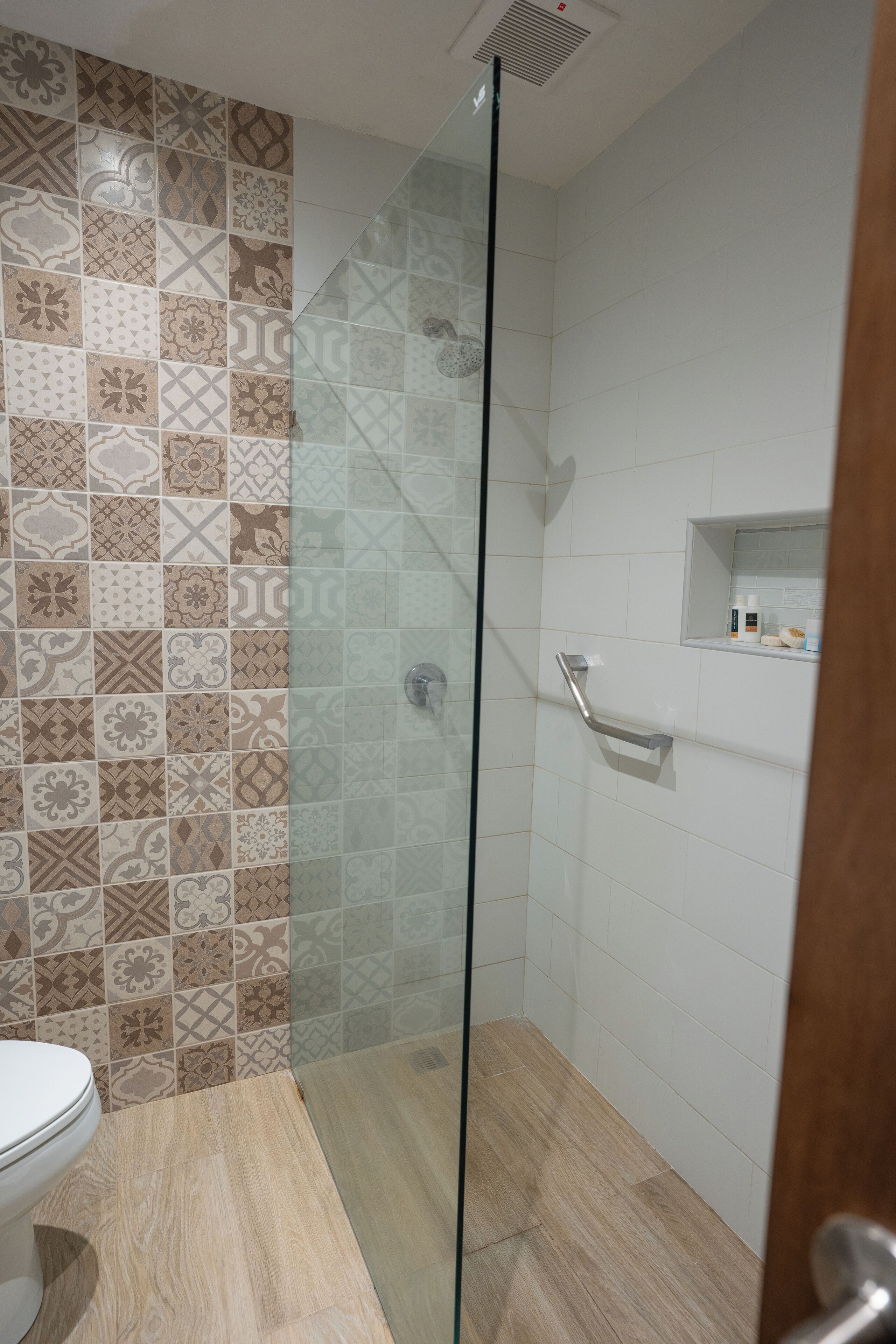 A bathroom with patterned tiles, a glass shower enclosure, a toilet, and a small shelf with toiletries.