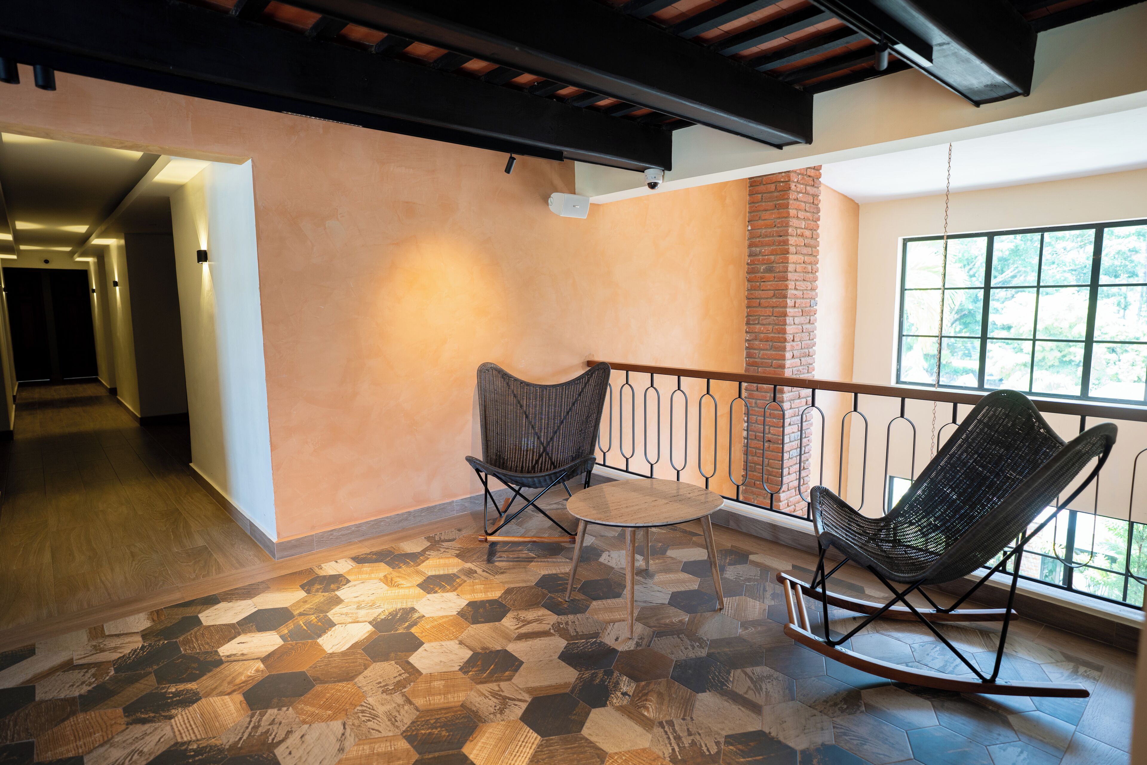 Una habitación amplia con suelo de patrón hexagonal, pared de ladrillo y una ventana grande que ofrece vistas a la vegetación. Hay dos mecedoras y una mesita.