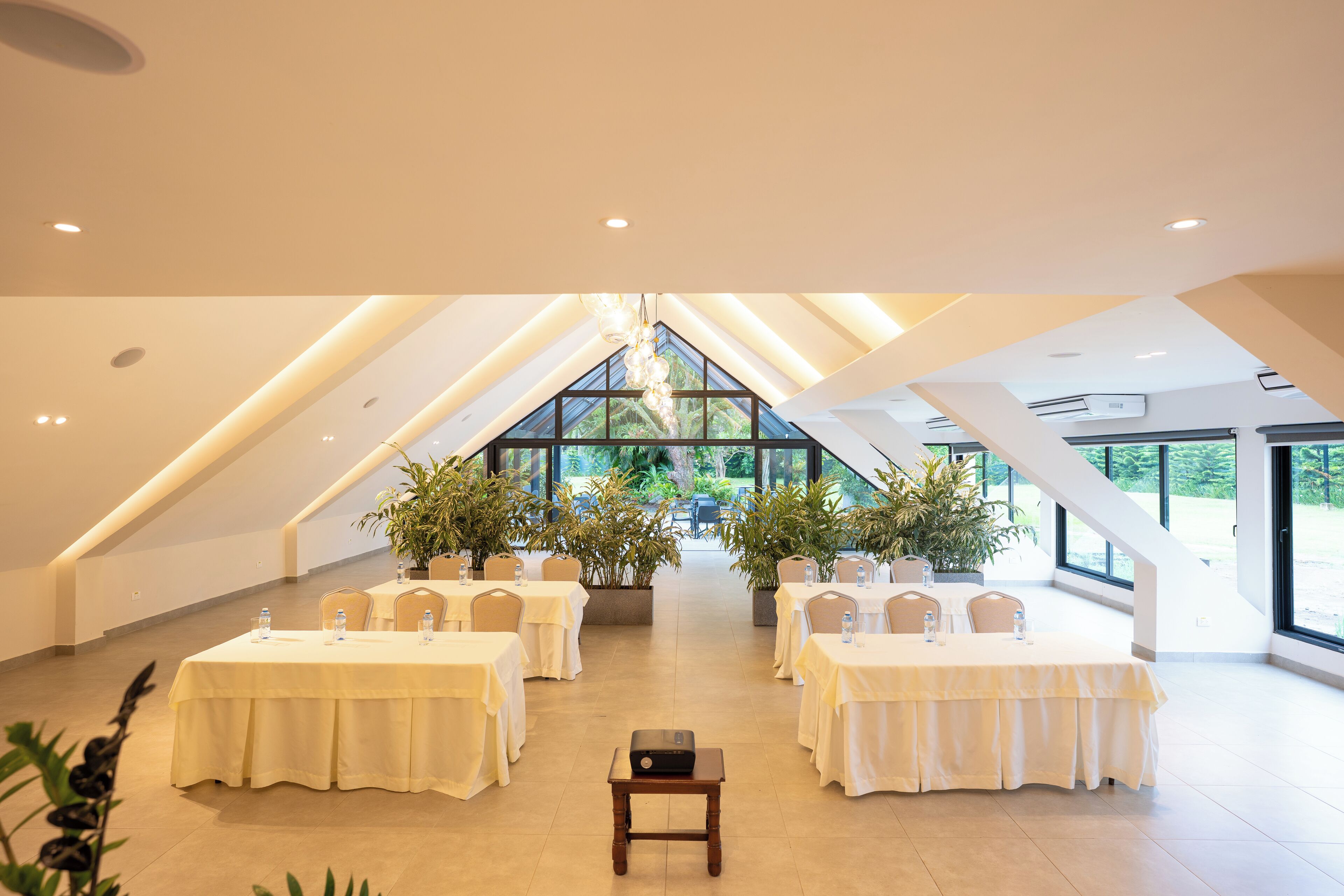 A spacious, well-lit conference room with high ceilings and large windows, featuring multiple tables and chairs, and potted plants.