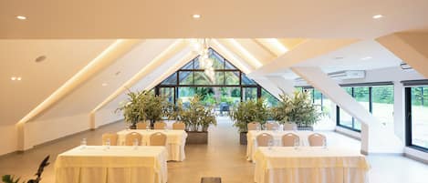 A spacious, well-lit conference room with high ceilings and large windows, featuring multiple tables and chairs, and potted plants.