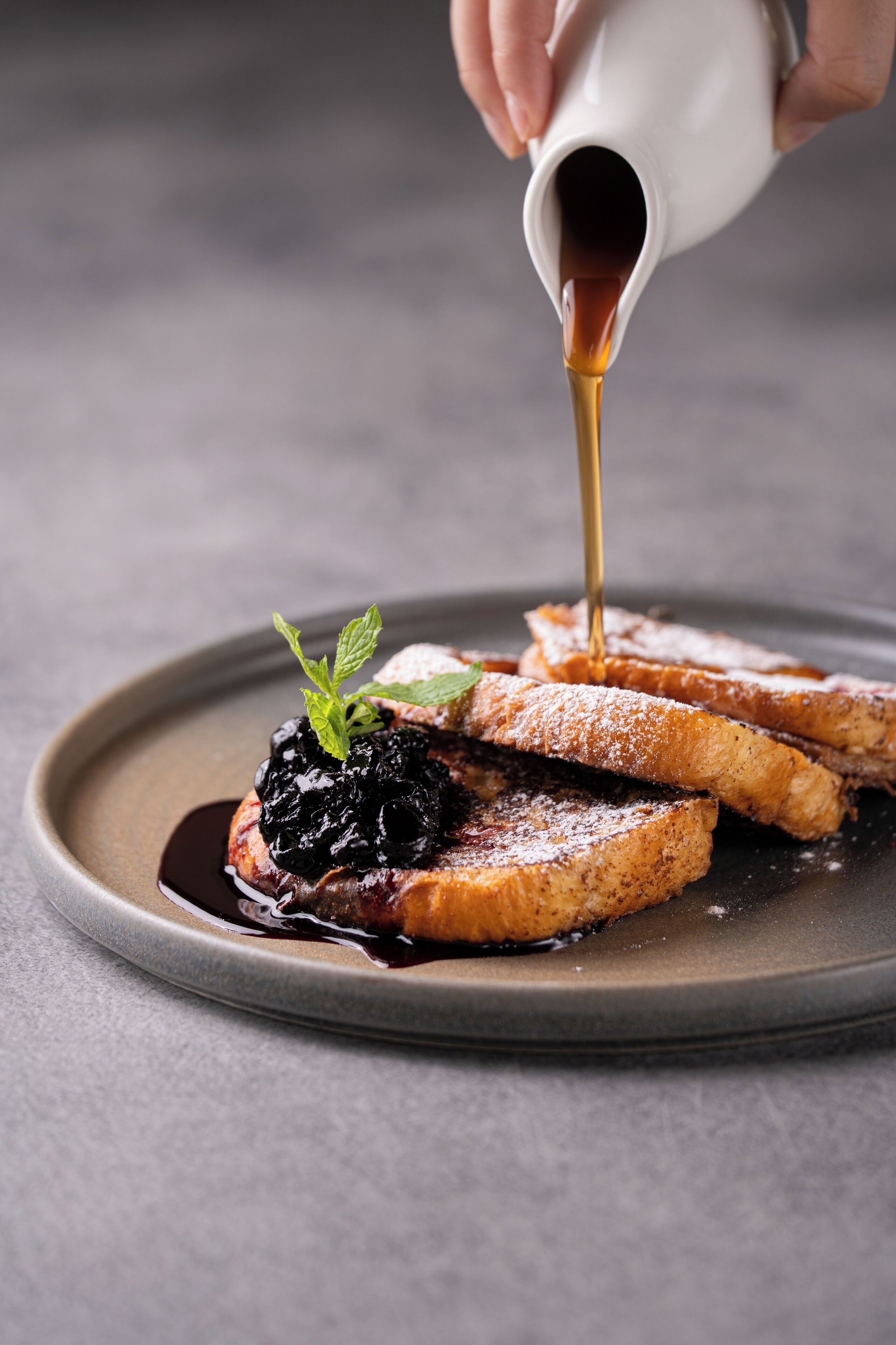 A plate with French toast topped with a berry compote and a mint leaf, being drizzled with syrup.