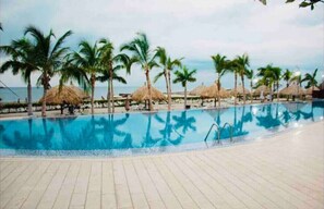 A swimming pool with palm trees and thatched umbrellas.