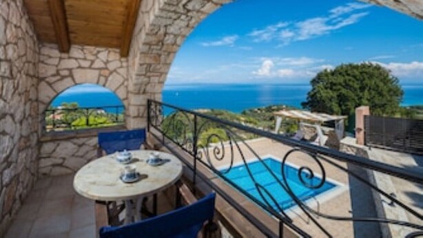 Un balcón con paredes de piedra y vistas al mar, una piscina y una mesa con sillas.