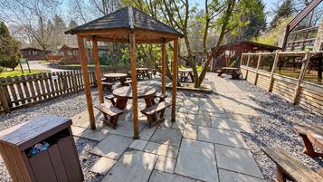 Un espace extérieur aménagé en terrasse, avec des tables de pique-nique, un gazebo en bois et une poubelle.