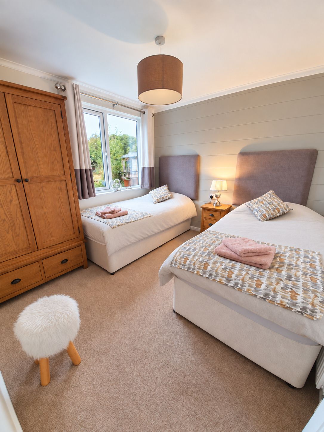 A bedroom with two beds, a wardrobe, a window with curtains, a lamp, and a fluffy stool.
