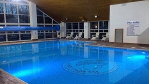 An indoor swimming pool with a clear view of the pool rules sign, lounge chairs, and a covered area with windows.