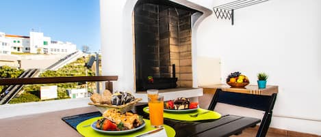Un balcon avec une table dressée pour deux personnes, où l’on peut déguster un repas composé de pain, de fruits et d’un verre de jus d’orange.