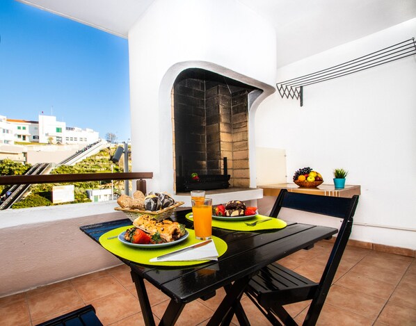 A balcony with a table set for two, featuring a meal with bread, fruit, and a glass of orange juice.