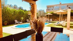 Zona de piscina amb una pèrgola de fusta, taula de vidre amb un ram d'estels secs i vista d'una piscina i un edifici al fons.
