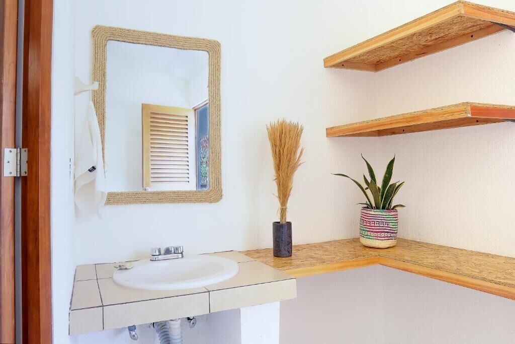 A bathroom with a white sink, a mirror with a woven frame, a potted plant, and wooden shelves.