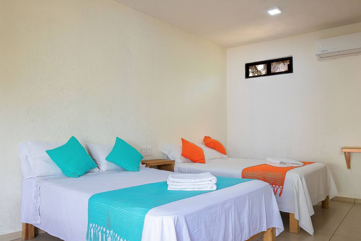 A hotel room with two beds, each with white bedding and colorful pillows, a small window, and an air conditioning unit on the wall.