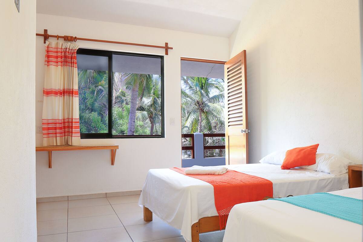 A bedroom with two beds, a window with a view of palm trees, and a balcony door.