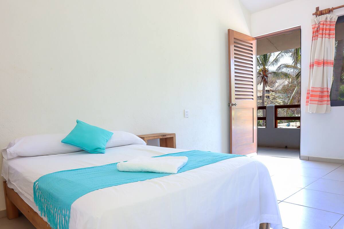 A neatly made bed with a blue throw blanket and pillows, a wooden headboard, and a view of a balcony with a door leading outside.