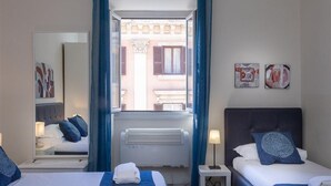 A hotel room with two beds, a window with blue curtains, a radiator, and artwork on the walls.