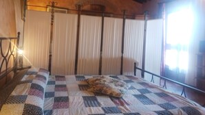 A bedroom with a checkered bedspread, a single pillow, a radiator, and a window with curtains. - Torre Prima 1 (San Gimignano)