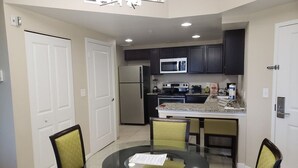 A compact kitchen with dark cabinets, a refrigerator, microwave, stove, and a dining area with a glass table and chairs. - 2 BR - Resort -2 Suite En Orlando Kissimmee, Cerca a los Parques Temáticos (Kissimmee)