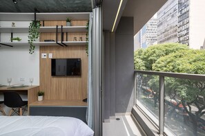 Una habitación de hotel moderna con cama, escritorio y silla, televisor montado en la pared y balcón con vista a la ciudad.