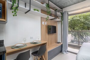 A modern hotel room with a dining area, a bed, a TV mounted on the wall, and a balcony with a view of trees.