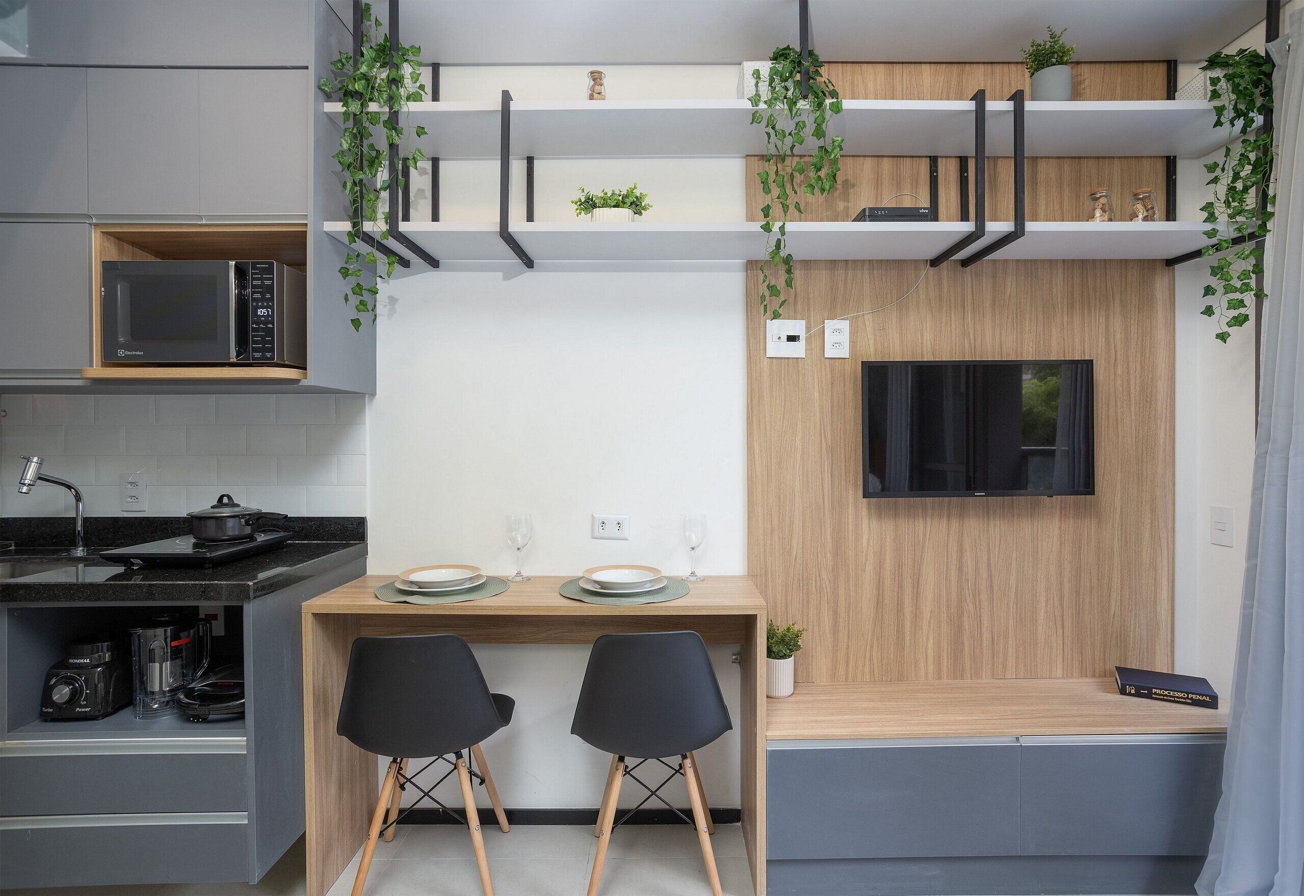 Cozinha compacta com micro-ondas, fogão e pia.  Possui área de refeições com duas cadeiras e uma mesa. Uma TV está fixada na parede.