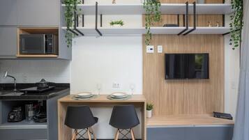 Cozinha compacta com micro-ondas, fogão e pia. Possui área de refeições com duas cadeiras e uma mesa. Uma TV está fixada na parede.