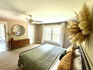 A bedroom with a bed, dresser, mirror, ceiling fan, and window with curtains.