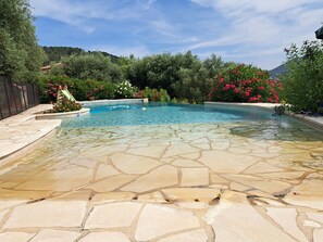 Piscina cristalina com um deck de pedra, rodeada por vegetação exuberante e plantas floridas.