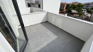 A balcony with grey tiles, a glass door, and a view of buildings and trees. - Sezer's Oliva Hotel (Ezine)